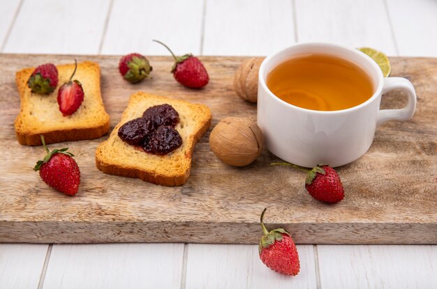 Vista dall'alto di deliziose fragole sul pane con una tazza di tè al lime su una tavola di cucina in legno su un fondo di legno bianco