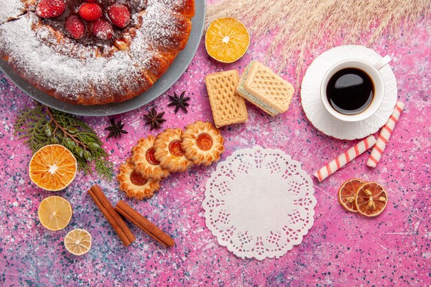 Вид сверху вкусный клубничный торт с печеньем, чаем и вафлями на светло-розовом фоне, выпечка сладкого сахарного бисквитного печенья, пирог
