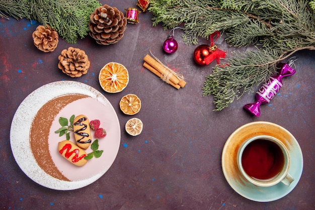 Вид сверху вкусного сладкого печенья с чашкой чая и рождественской елкой на темном фоне печенья сладкого печенья сахарного цвета
