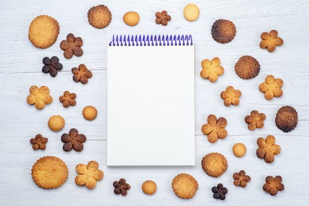 Top view of delicious sweet cookies different formed along with notepad on light desk, cookie biscuit sweet sugar