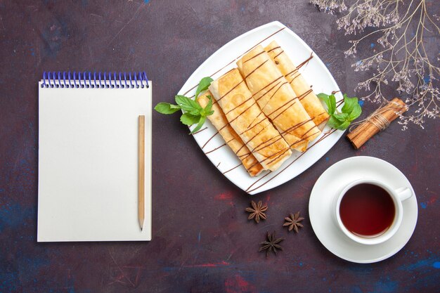 Top view delicious sweet pastries with cup of tea and notepad on dark space