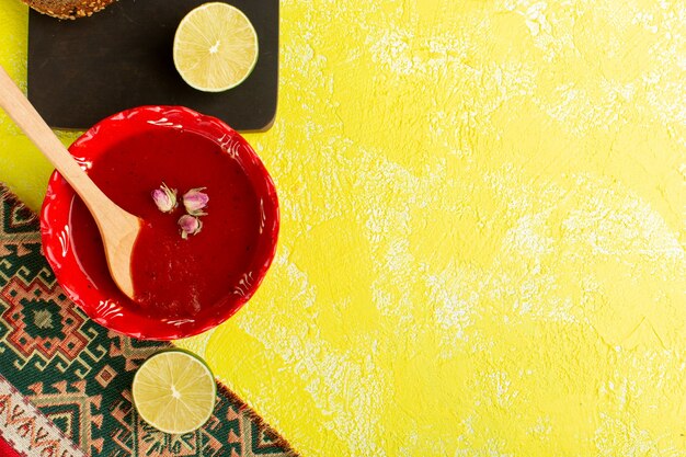 Вид сверху вкусный томатный суп внутри красной тарелки с лимонами на желтом столе, суп, ужин, овощная еда
