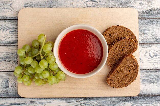 Top view delicious tomato soup with green grapes and dark bread loafs on grey table, soup food meal dinner