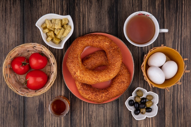 Top view of delicious turkish sesame bagels on a plate with olives on a bowl with tomatoes on a bucket and with a cup of tea on a wooden background