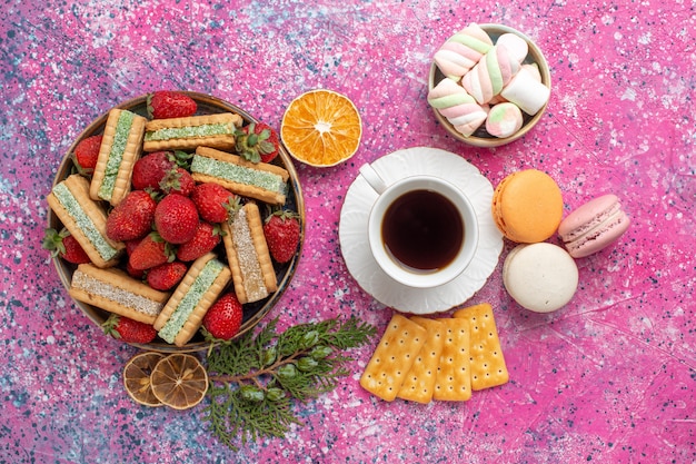 Top view of delicious waffles with cup of tea macarons and fresh red strawberries on pink surface