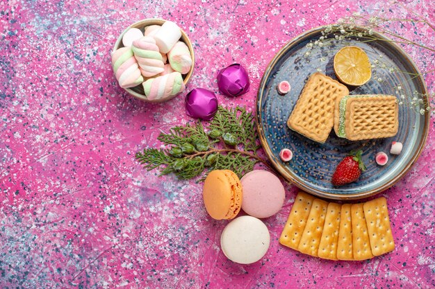 Top view of delicious waffles with french macarons and crackers on pink surface