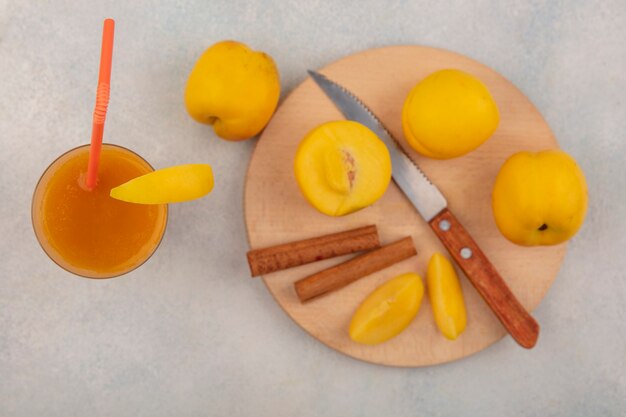 Вид сверху вкусных желтых персиков на деревянной кухонной доске с палочками корицы с ножом со свежим персиковым соком на стакане на белом фоне