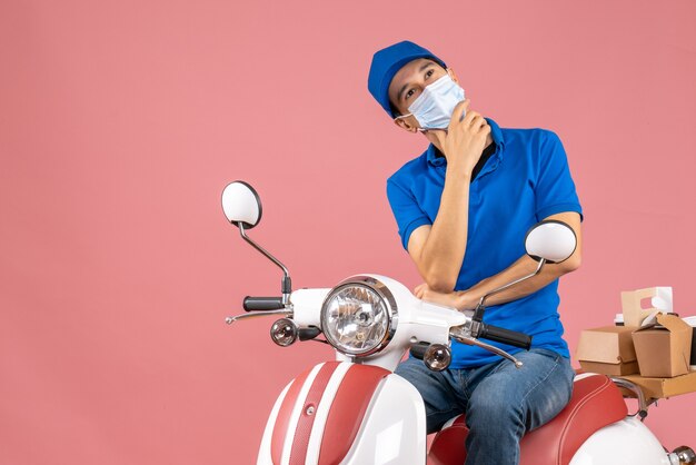 Top view of delivery guy in medical mask wearing hat sitting on scooter in deep thoughts on pastel peach background