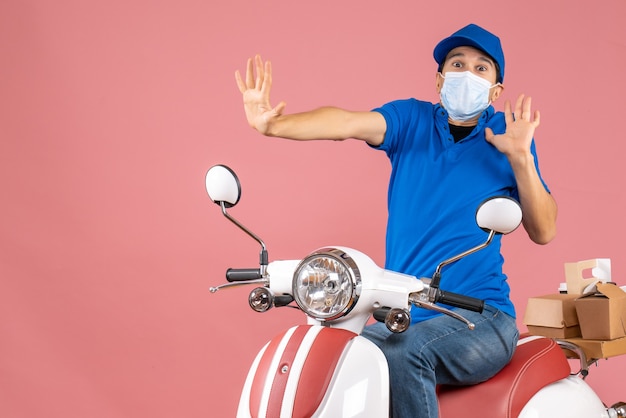 Top view of delivery guy in medical mask wearing hat sitting on scooter and feeling scared on pastel peach background