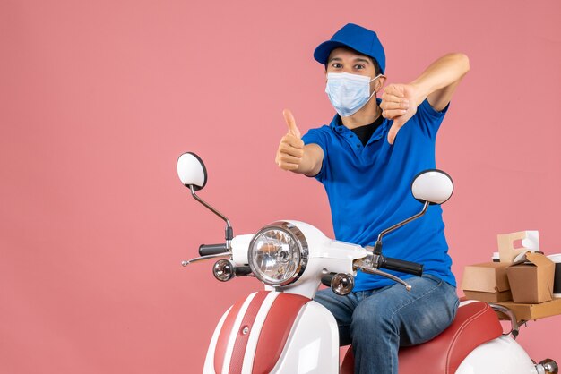 Top view of delivery guy thumbs up and thumbs down in medical mask wearing hat sitting on scooter on pastel peach background