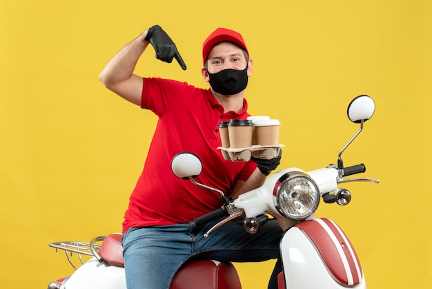 Top view of delivery guy wearing uniform and hat gloves in medical mask sitting on scooter showing orders feeling confident