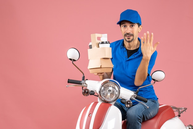 Top view of determined courier man wearing hat sitting on scooter holding orders showing five on pastel peach background