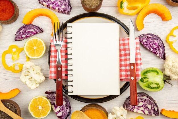 丸いプレートカット野菜のメモ帳フォークとナイフに書かれた上面図ダイエット白い木製のテーブルのボウルにさまざまなスパイス
