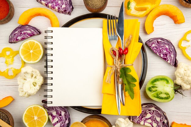Top view diet written on notepad tied fork and knife on yellow napkin on round plate cut vegetables different spices in bowls on white table