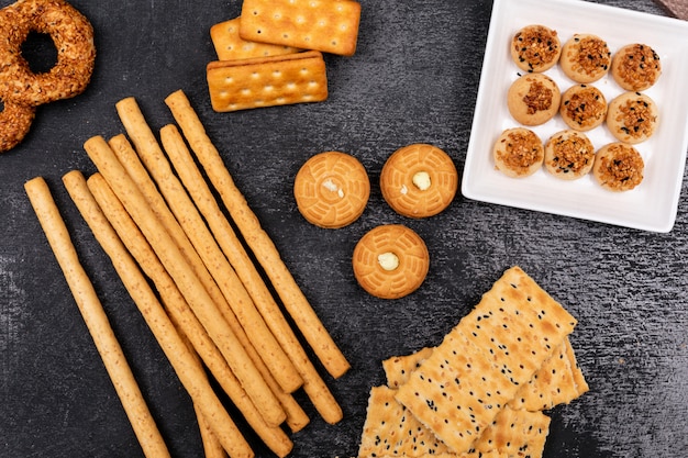 top view different cookies and bread sticks on dark surface