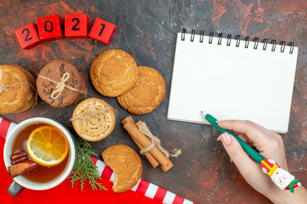 Top view different cookies cup of tea cinnamon sticks wood blocks notepad green pen in female hand on dark red table