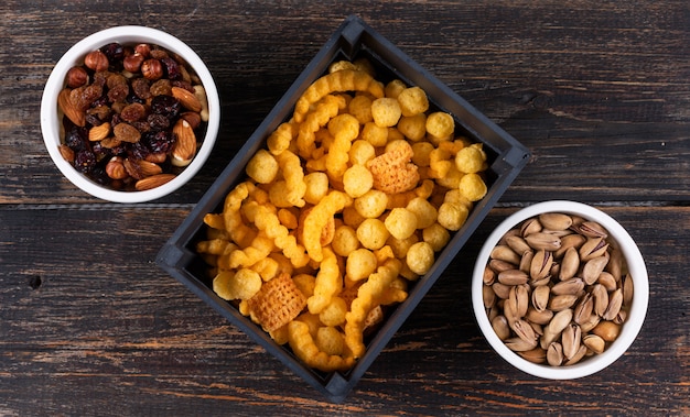 Top view of different kind of snacks as corn balls in black crate and nuts with dried fruits, pistachios in bowls on dark wooden surface horizontal