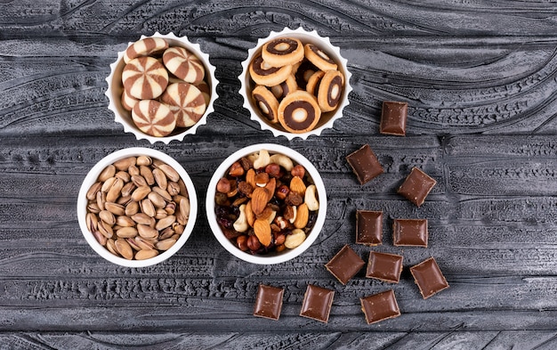 Вид сверху различных видов закусок, таких как орехи, печенье и шоколад в мисках на темной поверхности горизонтальной