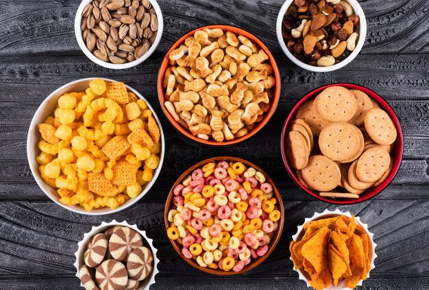 Top view of different kind of snacks as nuts, crackers and cookies in bowls on dark surface horizontal