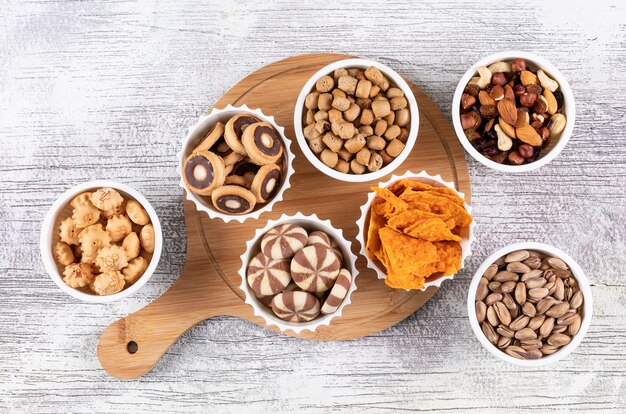 Top view of different kind of snacks as nuts, crackers and cookies in bowls on wooden cutting board on white surface horizontal