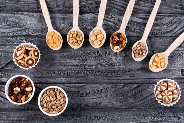 Top view of different kind of snacks as nuts and crackers on wooden spoons and cookies in bowls with copy space on dark surface horizontal