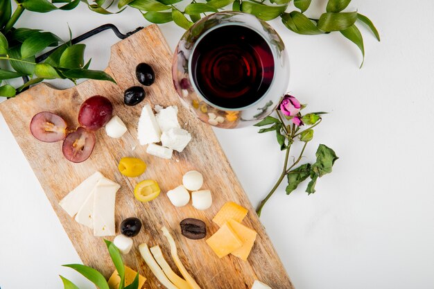 Top view of different kinds of cheese with grape pieces olives on cutting board with red wine on white decorated with flowers and leaves with copy space 1