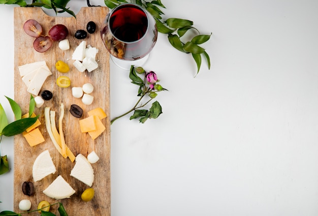 Вид сверху различных видов сыра с кусочками винограда оливки на разделочной доске с красным вином на белом украшенные цветами и листьями с копией пространства
