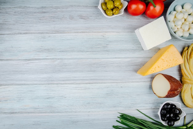 Top view of different kinds of cheese with green onion, fresh tomatoes and pickled olives on grey wooden table with copy space