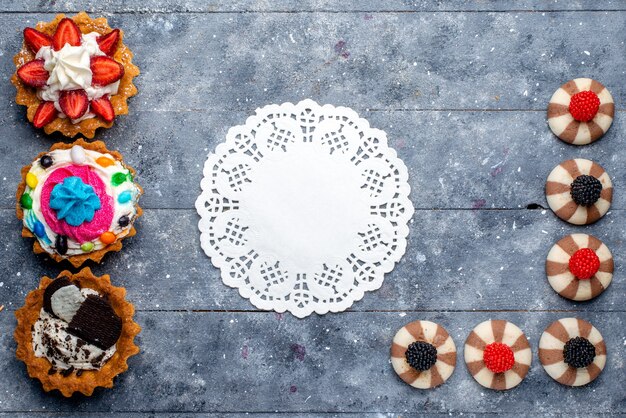 Top view of different little cakes with sliced fruits candies chocolate and berries on grey desk, cookie biscuit sweet sugar bake