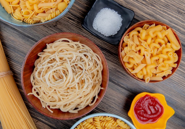 Top view of different macaronis as spaghetti rotini vermicelli and others with salt and ketchup on wood