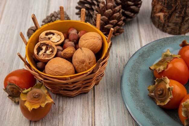 Top view of different nuts on a bucket with soft fresh persimmons on a plate on a grey wooden wall