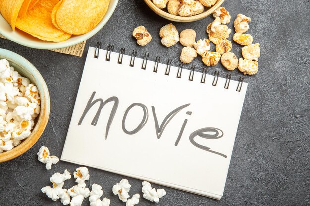 Top view different snacks with movie written notepad on dark background