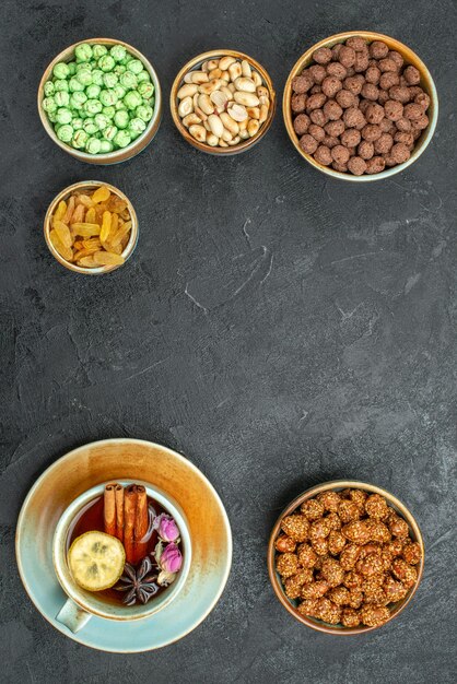 Top view of different sweet candies with nuts and cup of tea on black grey