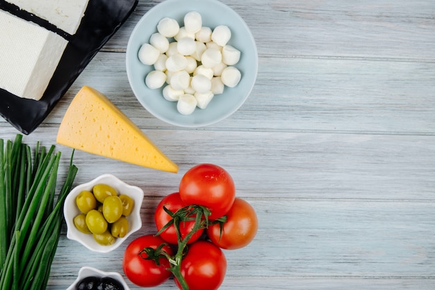 Top view of different types of cheese with fresh tomatoes and pickled olives on grey wooden table with copy space