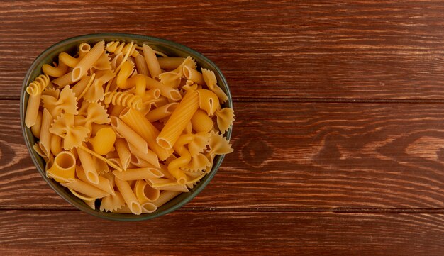 Top view of different types of macaroni in bowl on wood with copy space