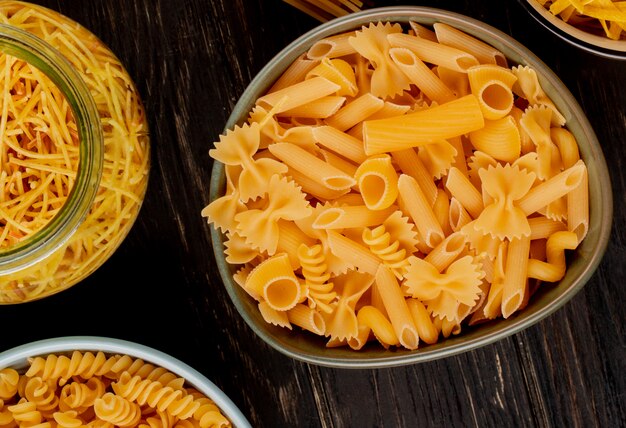 Top view of different types of pasta as spaghetti rotini and other types in bowls and jar on wooden surface
