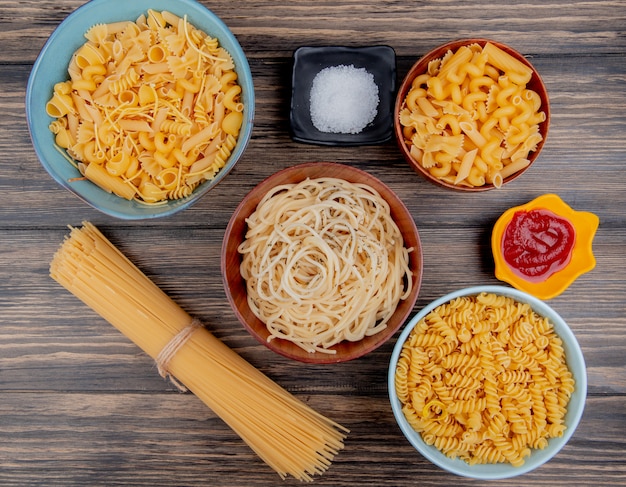 Top view of different types of pasta as spaghetti rotini vermicelli and others with salt and ketchup on wooden surface