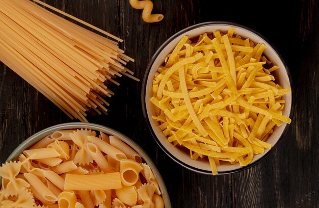 Top view of different types of pasta as tagliatelle and other ones in bowls with spaghetti type on wooden surface