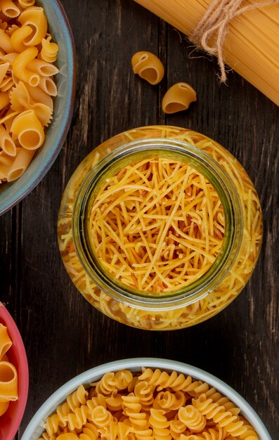 Top view of different types of pasta as vermicelli rotini and others in jar and bowls on wooden surface