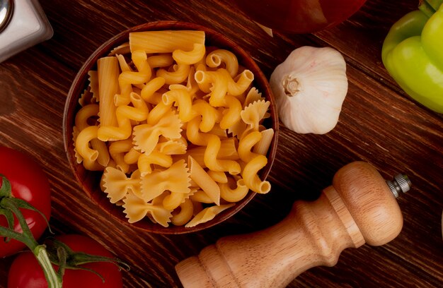 Top view of different types of pasta in bowl with salt tomato garlic pepper on wooden surface