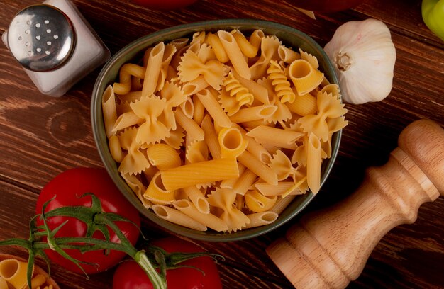 Top view of different types of pasta in bowl with salt tomato garlic on wooden surface