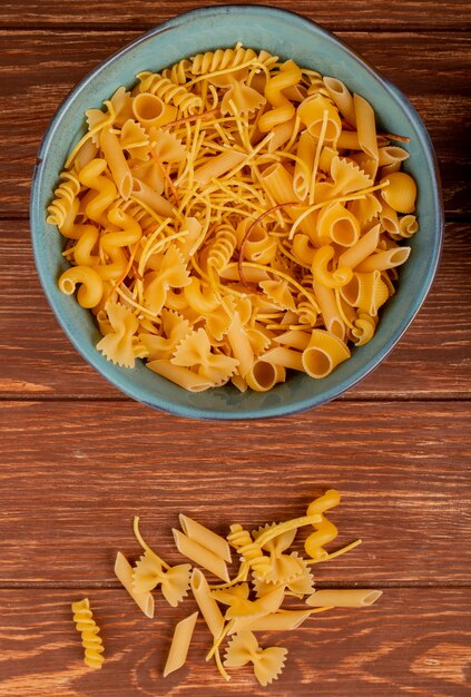 Top view of different types of pasta in bowl and on wooden surface