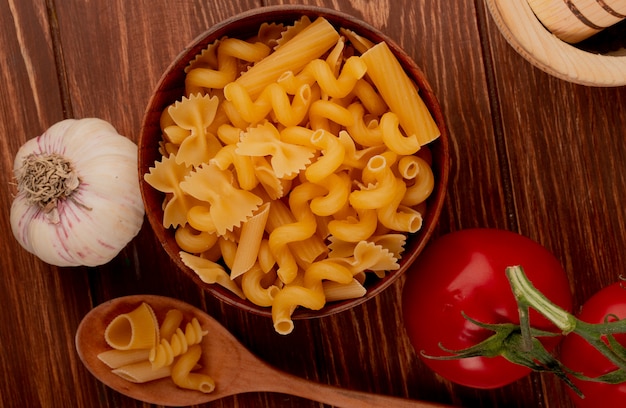 Top view of different types of pasta in wooden bowl and spoon with tomato and garlic on wooden surface