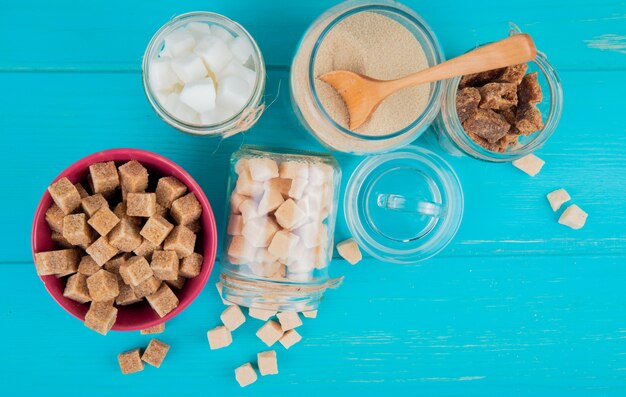 Top view of different types of sugar in bowls and in glass jars on blue wooden background