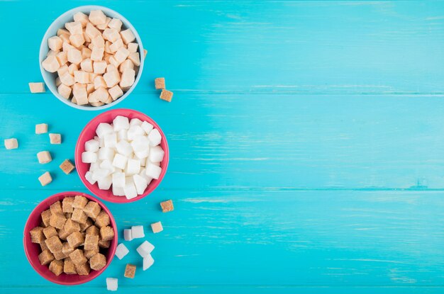 Top view of different types of sugar cubes in bowls on blue wooden background with copy space