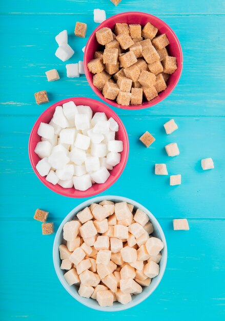 Top view of different types of sugar cubes in bowls on blue wooden background