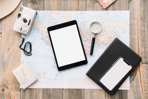 Top view of digital tablet; cell phone; magnifying glass and diary on map against wooden background