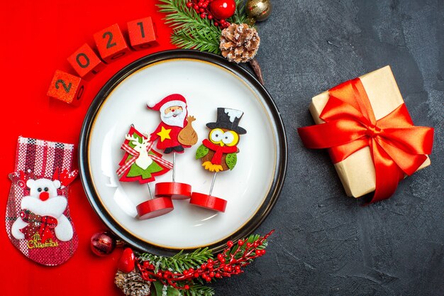 Top view of dinner plate decoration accessories fir branches and numbers christmas sock on a red napkin next to gift on a black table