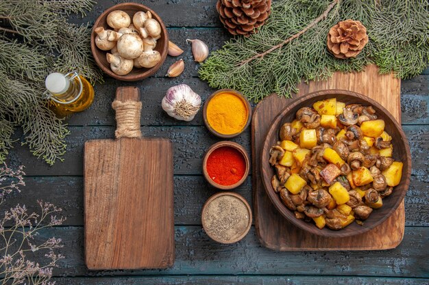 Вид сверху блюдо и доска с грибами и картофелем на деревянной доске рядом с красочными специями, разделочная доска, масло в бутылке, чеснок, миска с грибами и ветки с шишками