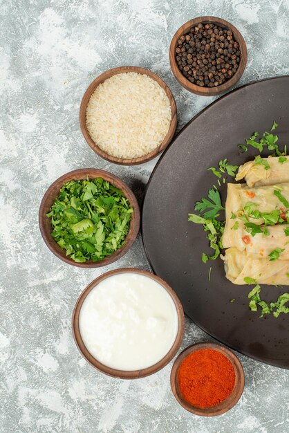 Top view dish in plate stuffed cabbage in plate next to the bowl with herbs rice colorful spices sour cream and black pepper on the right side of grey table